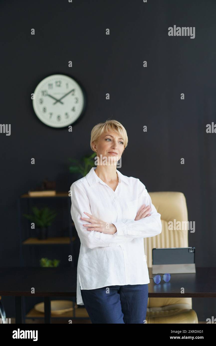 Una donna sta in piedi con le braccia incrociate, pensando alla sua prossima mossa in un ufficio elegante pieno di luce naturale e di un grande orologio a parete. Foto Stock