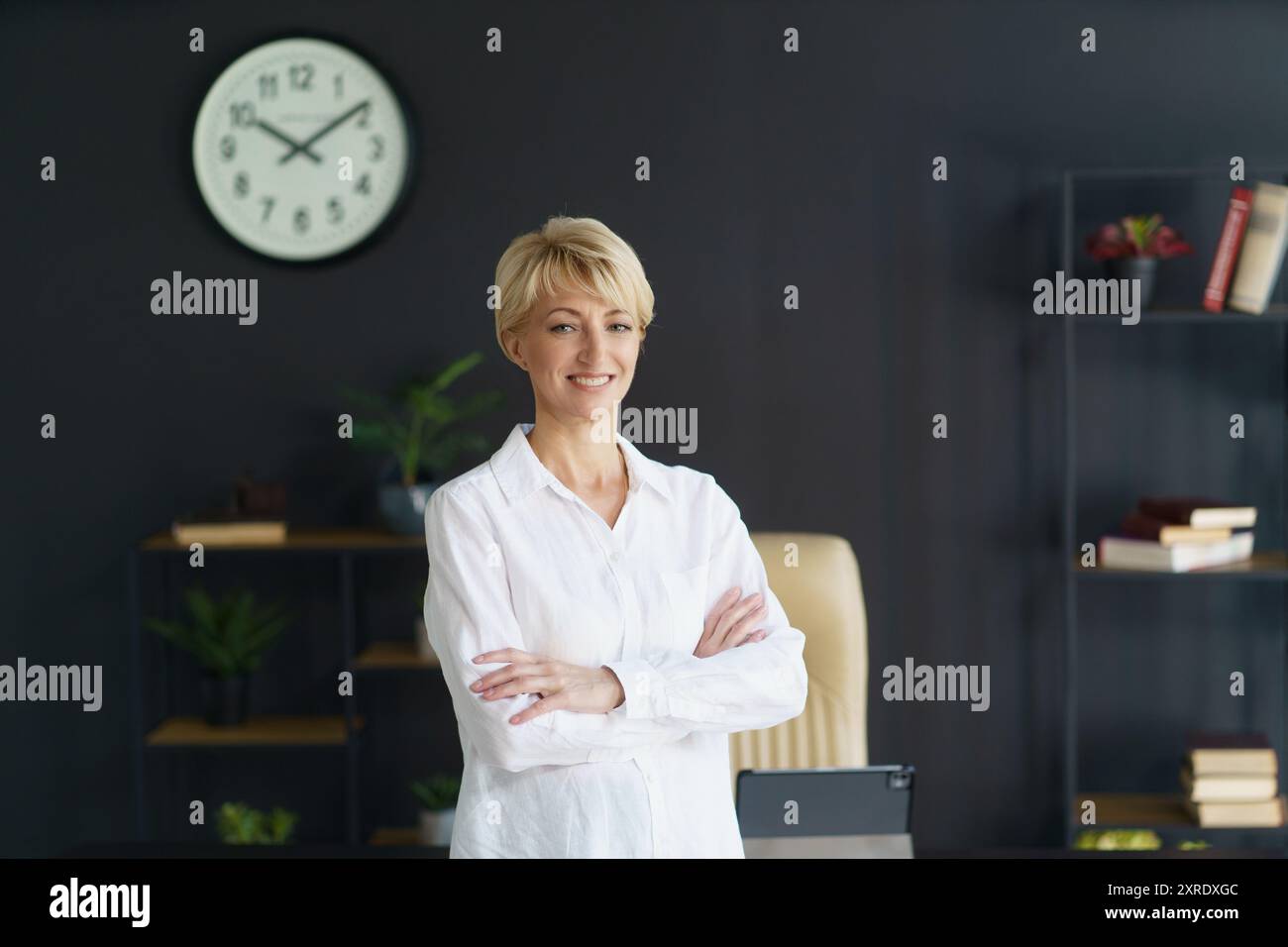 Una donna pronta con un sorriso sicuro si trova in un ufficio elegante, irradiando professionalità mentre l'orologio mostra l'ora sullo sfondo. Foto Stock