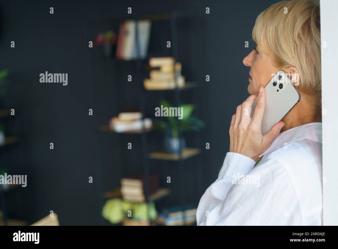 Una donna immersa nella conversazione sul suo smartphone, circondata da scaffali di libri e vegetazione lussureggiante in una stanza elegante. Foto Stock