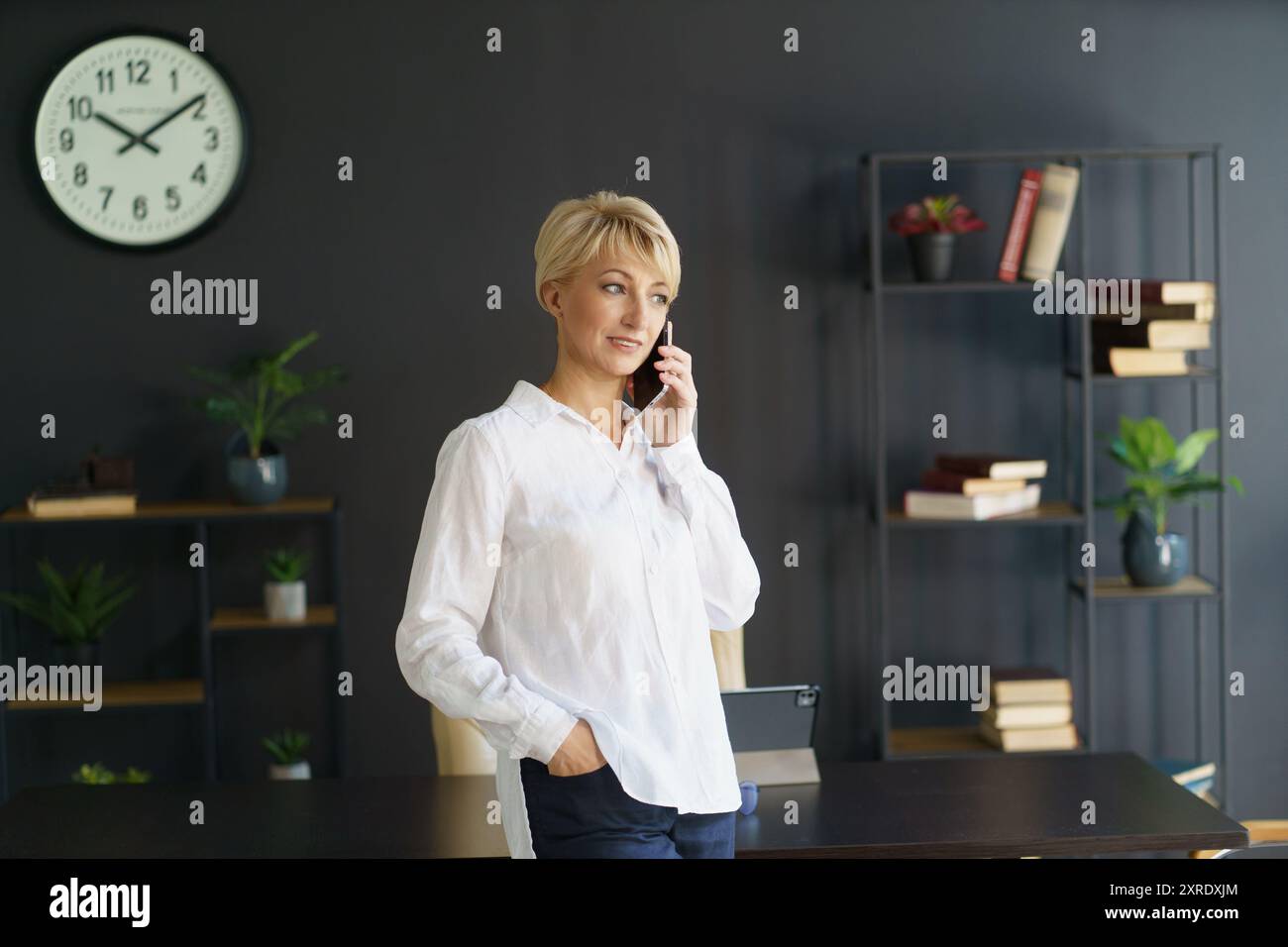 Una donna sicura di sé si trova in un ufficio elegante, conversando al telefono, circondato da libri e vegetazione, trasudando un senso di professionalità e di tranq Foto Stock