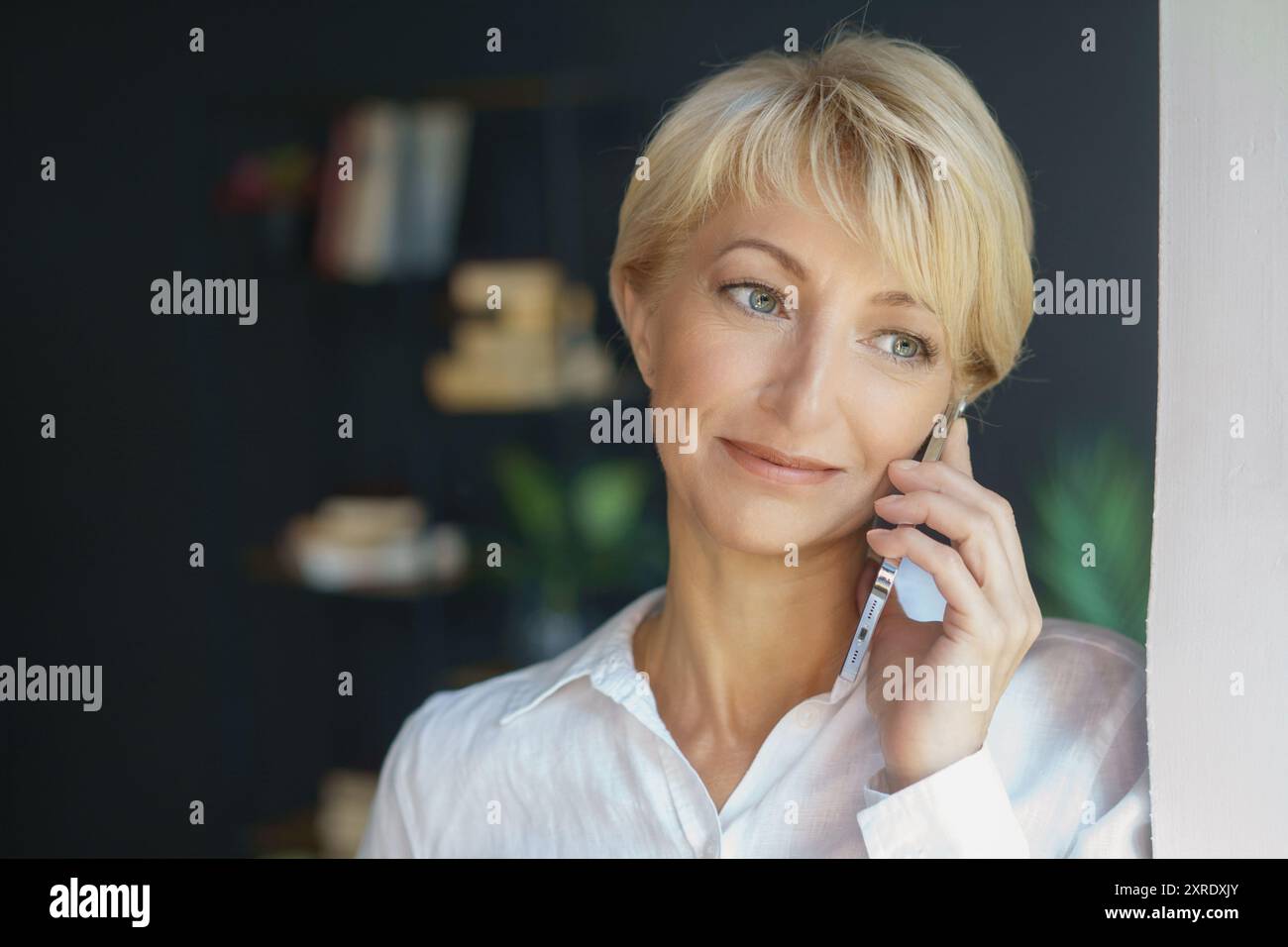 Una donna con una camicetta bianca sorride mentre parla al telefono, trasmettendo calore e coinvolgimento in un ambiente splendidamente accentuato. Foto Stock