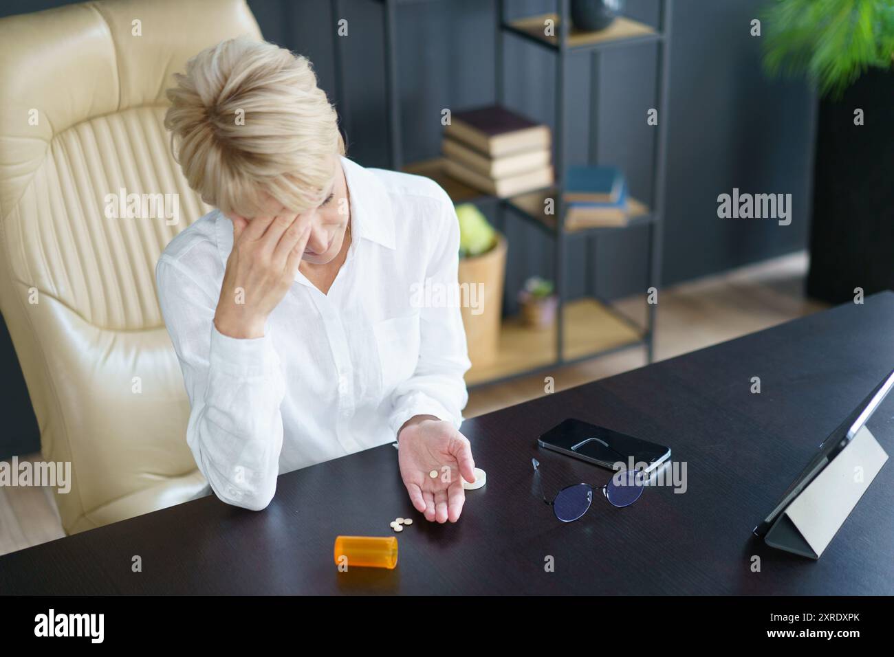 Una donna in difficoltà siede alla sua scrivania, tenendo le medicine in mano mentre riflette sui suoi pensieri travolgenti in un ufficio contemporaneo. Foto Stock