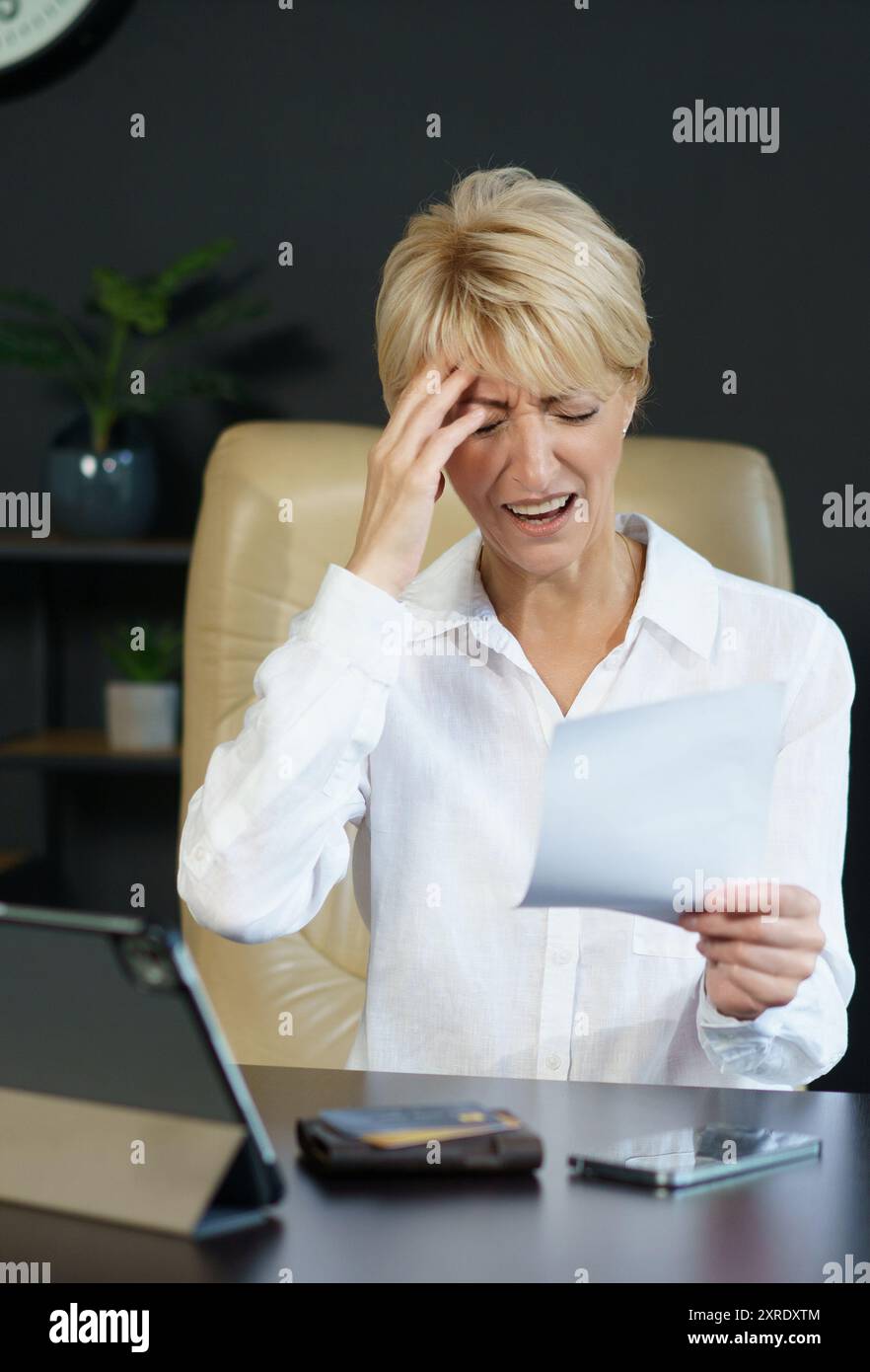 Una donna siede alla sua scrivania, visibilmente angosciata mentre esamina un documento, circondata da elementi essenziali dell'ufficio e da una pianta in vaso. Foto Stock Una donna siede alla sua scrivania, visibilmente angosciata mentre esamina un documento, circondata da elementi essenziali dell'ufficio e da una pianta in vaso. Foto Stock