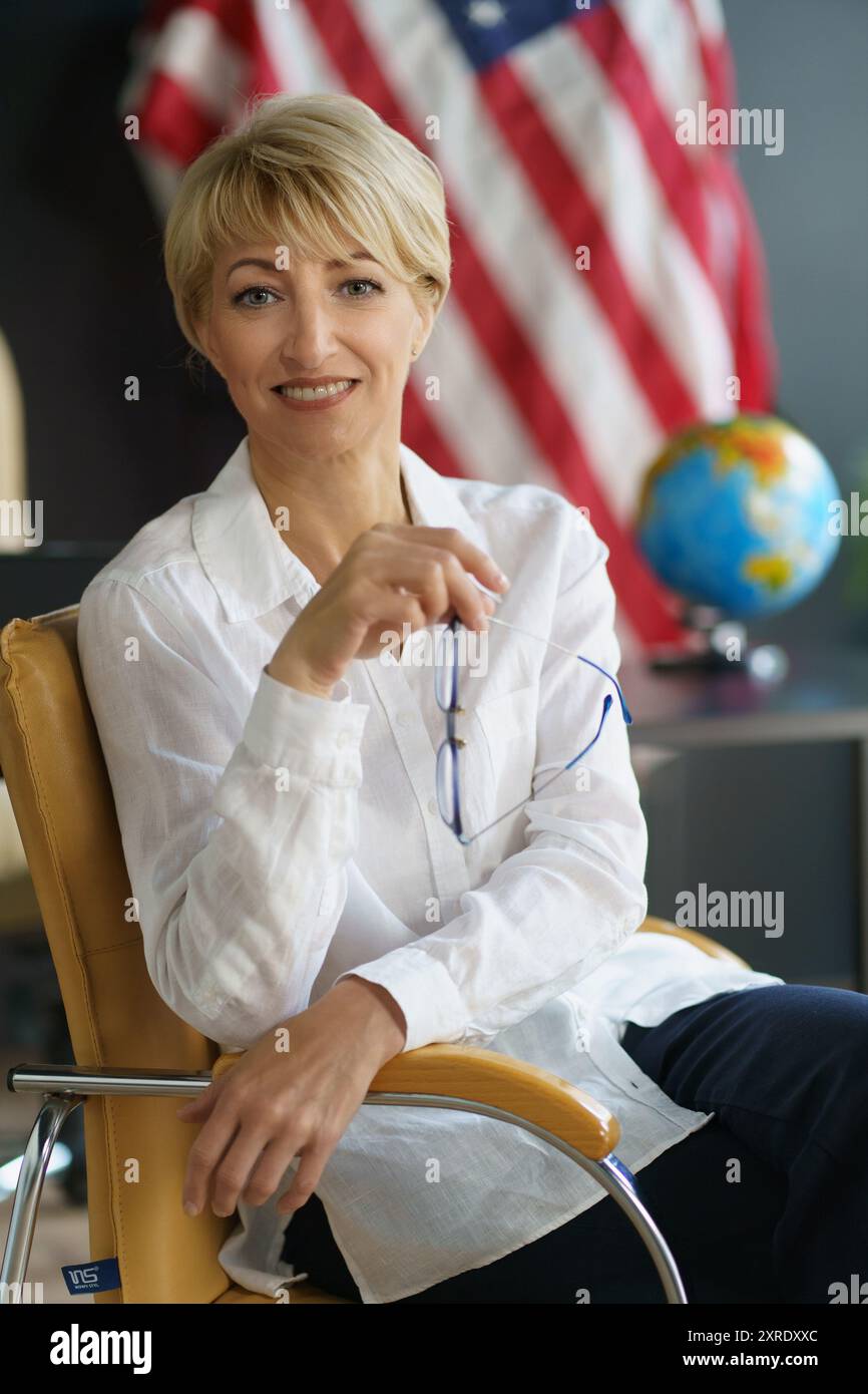 Un individuo sorridente si rilassa su una sedia da ufficio, tenendo gli occhiali, con una bandiera americana e globo sullo sfondo, incarnando un senso di professionalita' Foto Stock