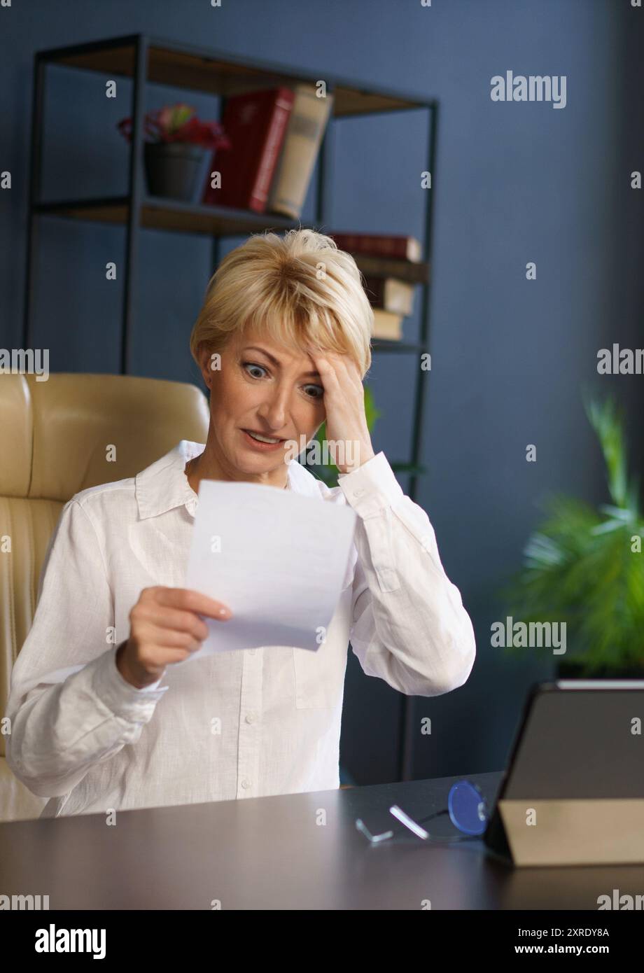 Una donna appare ansiosa mentre legge un documento, seduto alla sua scrivania con un tablet elettronico nelle vicinanze in un ambiente di ufficio contemporaneo. Foto Stock