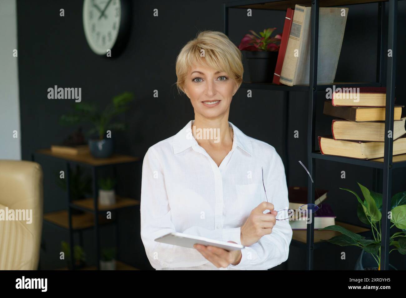 La donna è in piedi con sicurezza con un notebook e occhiali, circondata da libri e piante in un ambiente luminoso da ufficio. Foto Stock