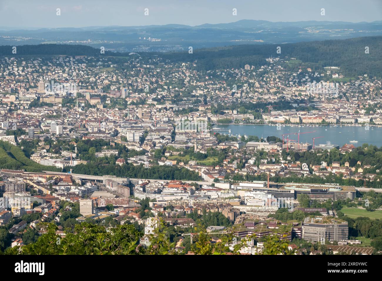 Panoramablick vom Uetliberg auf Zürich und den Zürichsee Das Bild zeigt einen weiten Panoramablick vom Uetliberg auf die Stadt Zürich, den Zürichsee und die umliegenden Stadtteile. Im Vordergrund ist die Autobahn A3 zu sehen, die sich durch die Stadt schlängelt und eine wichtige Verkehrsverbindung in der Region darstellt. Die Autobahn verbindet den südlichen Teil Zürichs mit anderen Teilen der Schweiz und verläuft hier durch das Stadtgebiet, vorbei an Büro- und Wohngebäuden. Weiter hinten breitet sich die dichte Bebauung Zürichs aus, mit einer Mischung aus Wohnhäusern, Geschäftszentren und His Foto Stock