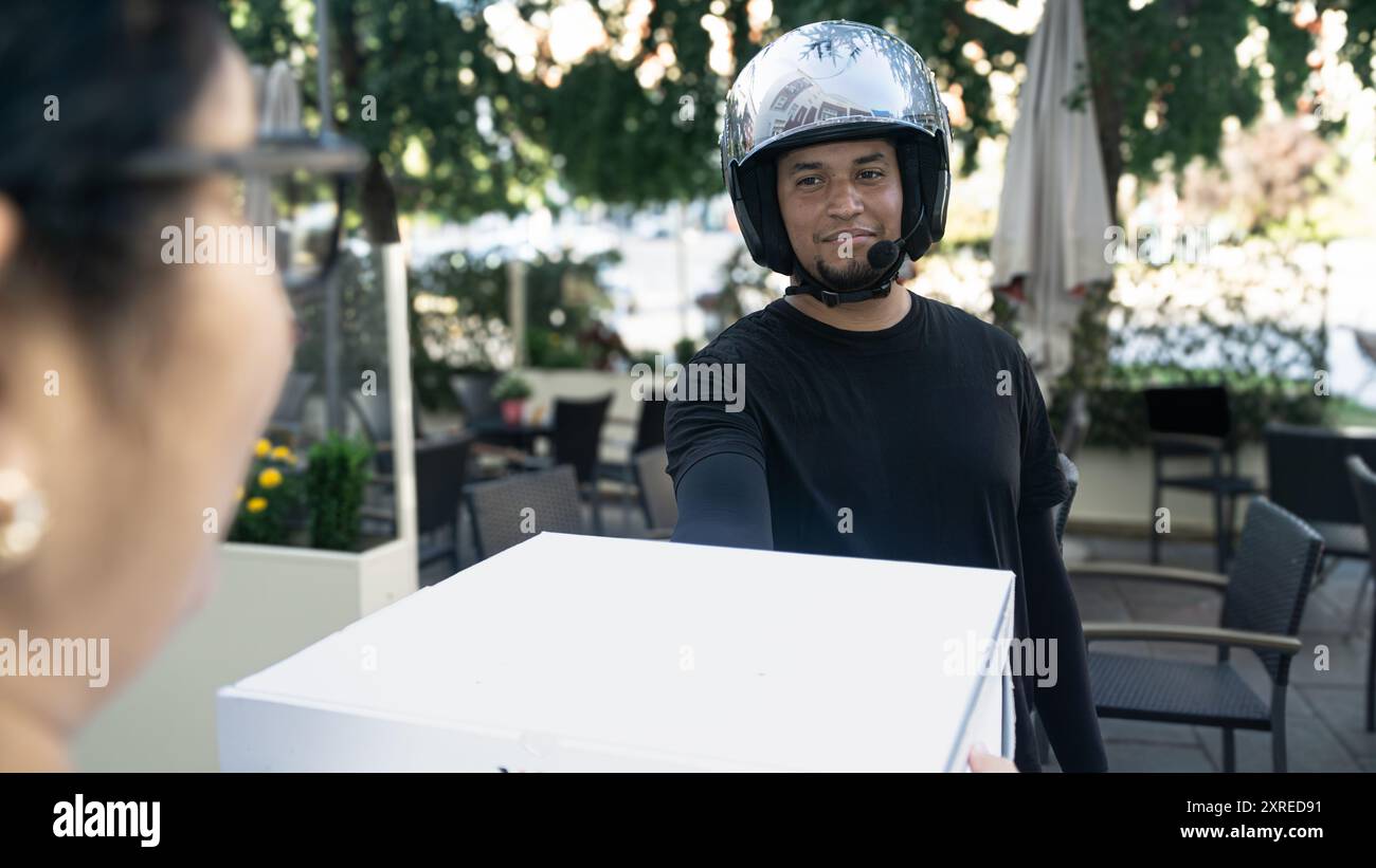 Un uomo che indossa un casco sta consegnando una scatola bianca a una donna. La scena è ambientata in un ristorante con diverse sedie e tavoli da pranzo Foto Stock