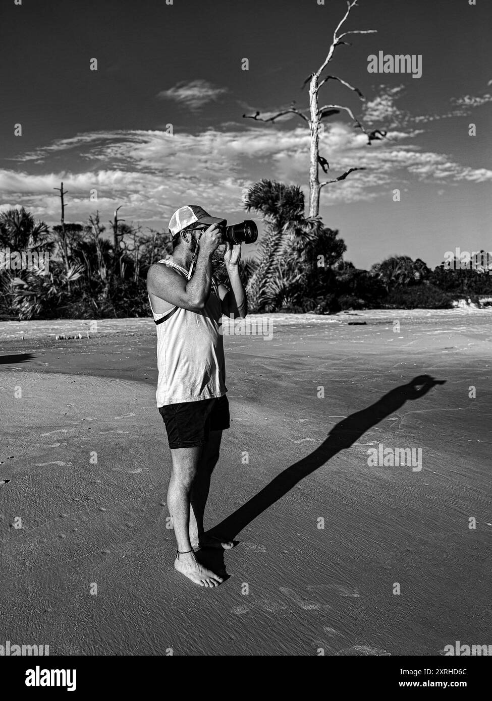 Attraente fotografo maschile che cattura il tramonto sulla spiaggia con abbigliamento casual e a piedi nudi Foto Stock