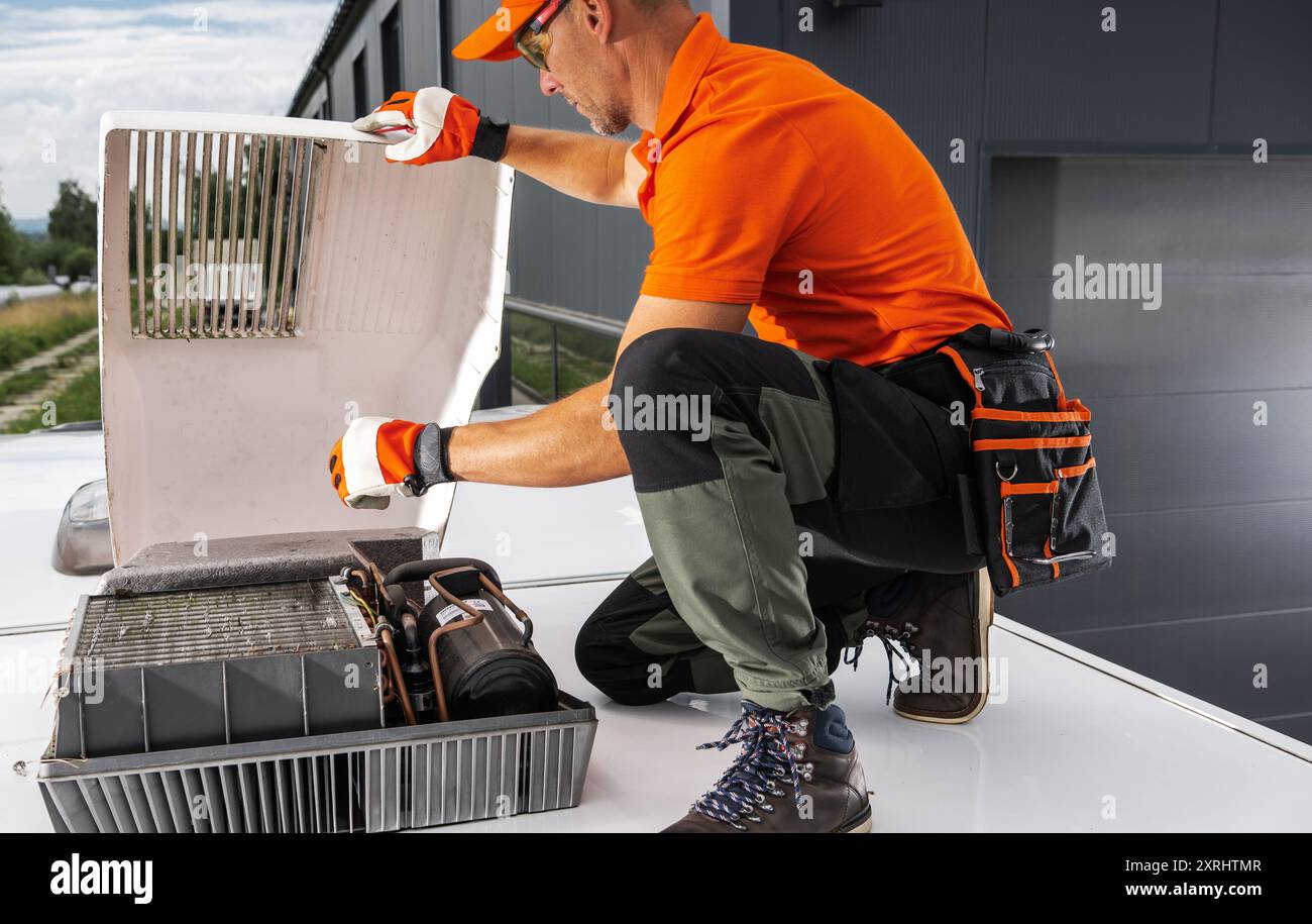 Un tecnico con una camicia arancione ispeziona e ripara un'unità HVAC sul tetto di un furgone camper indossando guanti e attrezzatura di sicurezza. Foto Stock