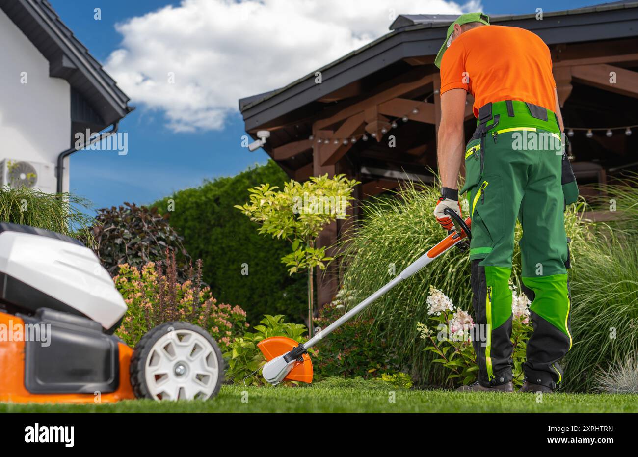 Un operaio in un abbigliamento vivace utilizza un rifinitore per mantenere il lussureggiante giardino, circondato da piante colorate sotto un cielo azzurro. Foto Stock