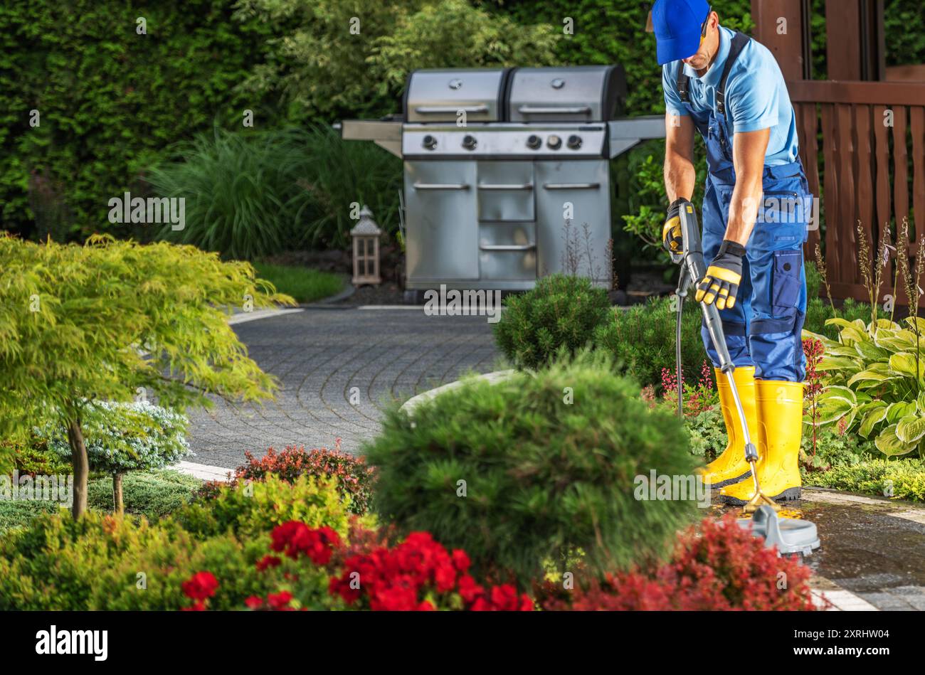 Un giardiniere con stivali gialli rifinisce i cespugli in un giardino splendidamente paesaggistico con fiori colorati e una griglia. Foto Stock