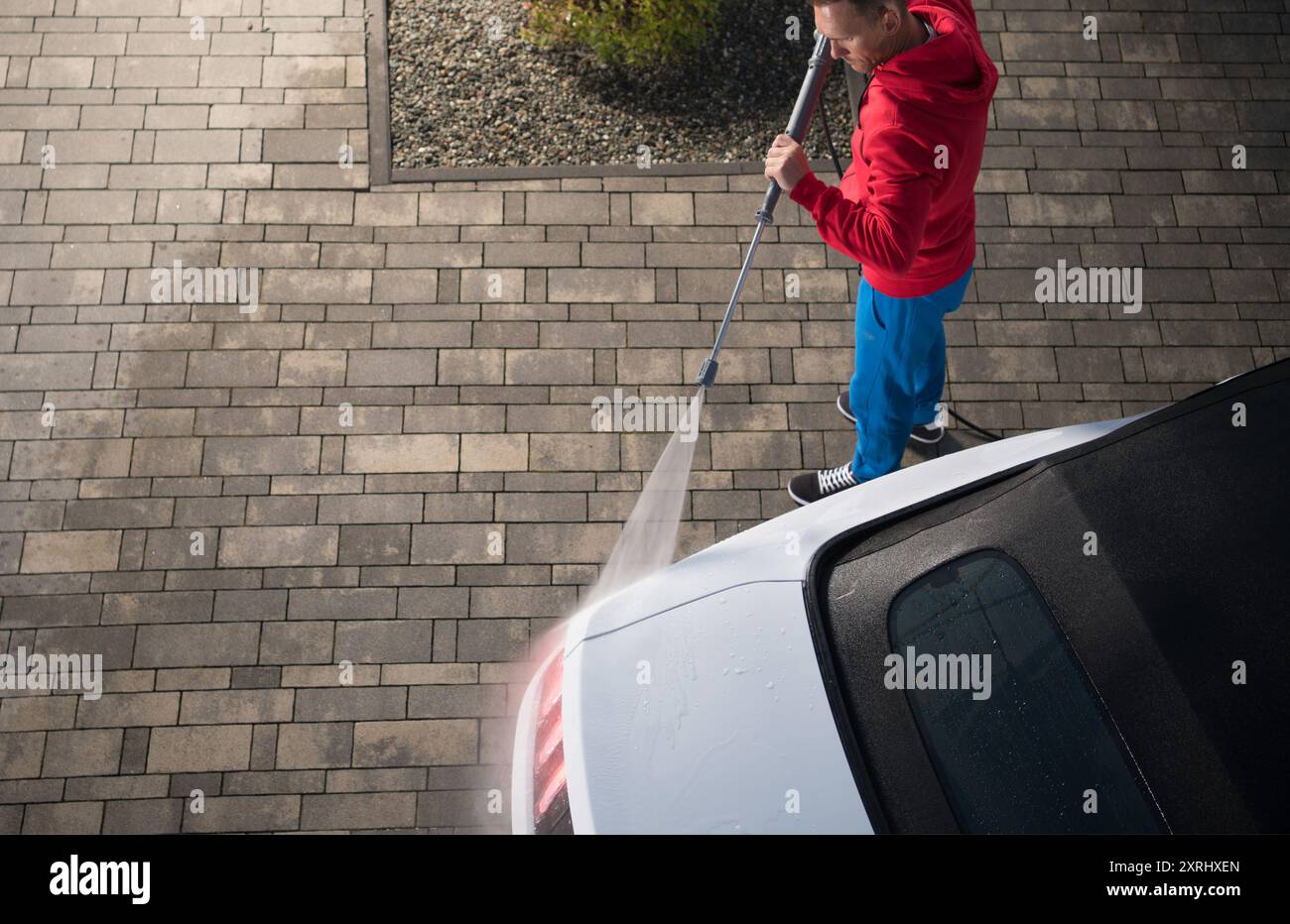 Una persona con cappuccio rosso e pantaloni blu utilizza una pulitrice a pressione per pulire un'auto parcheggiata in un vialetto, circondata da marciapiedi in pietra. Foto Stock