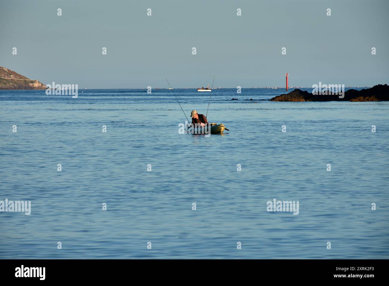 Un pescatore pesca tranquillamente da una canoa ricreativa nelle acque con la città di Bayonne sullo sfondo. Le acque calme forniscono un ambiente sereno Foto Stock