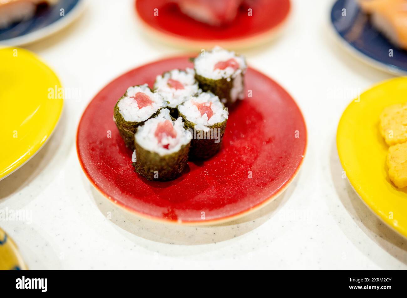 Primo piano di involtini di sushi al tonno maki preparati su un piatto rosso vivace, circondati da altri piatti di sushi su piatti colorati, che mettono in risalto una varietà Foto Stock