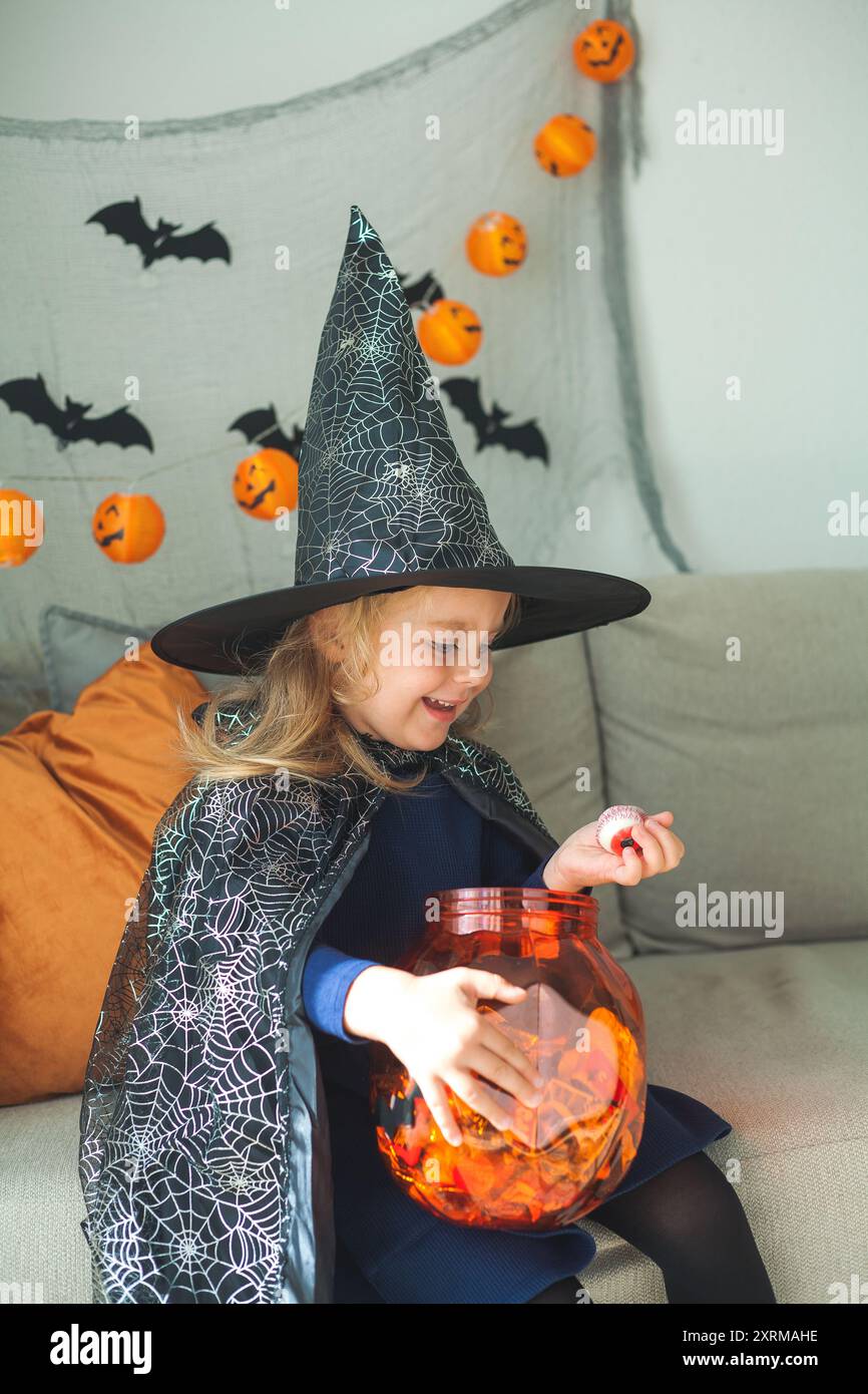 Una bambina di 3 anni in costume da strega con un secchiello di zucca pieno di dolci celebra Halloween a casa con decorazioni. Vacanze. Foto Stock