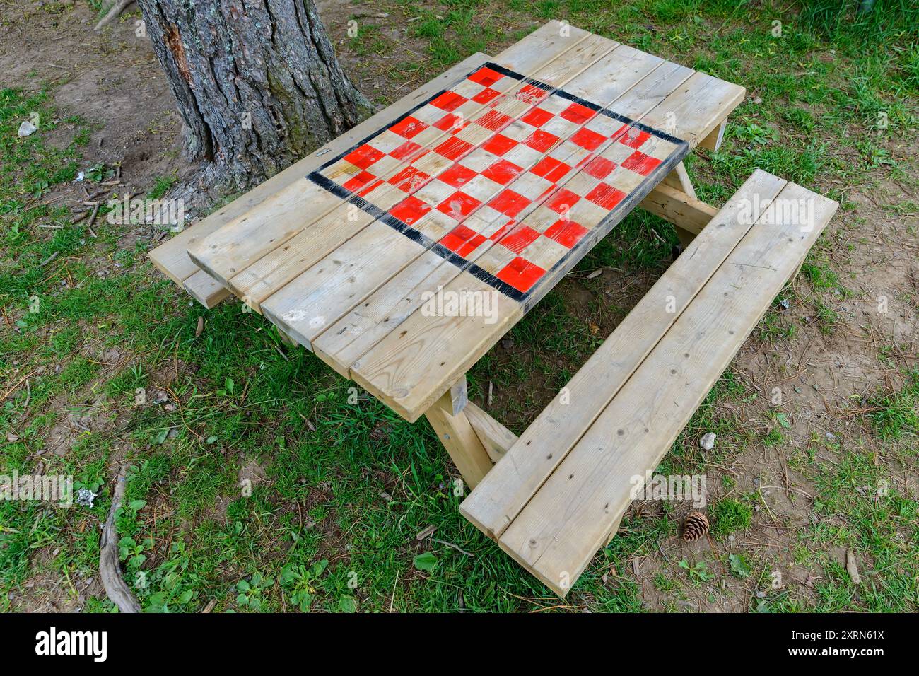 Un tavolo da picnic con scacchi o dama dipinta sulla parte superiore. Sembra vecchio e sbiadito. Si trova accanto a un tronco di albero e poggia su erba e sporco. Foto Stock