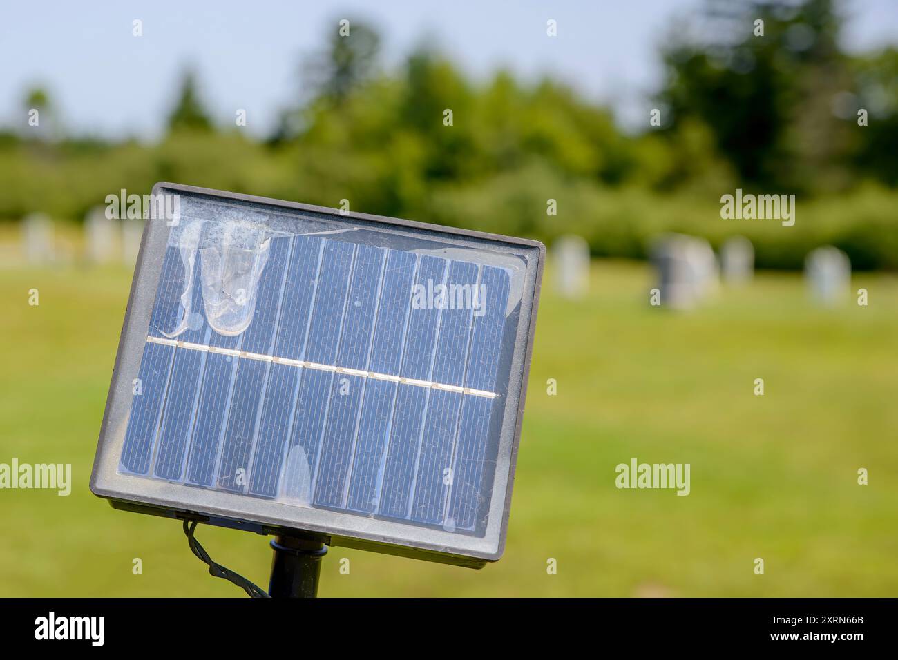 Un piccolo pannello solare danneggiato si attacca su un palo. Bassa profondità di campo, alberi e un piccolo cimitero dietro. Foto Stock