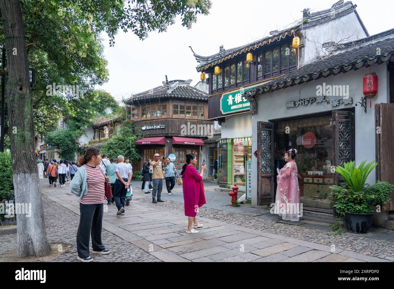 Suzhou, Cina - 11 giugno 2024: Una strada acciottolata fiancheggiata da edifici e negozi tradizionali cinesi, con gente che cammina nel verde. Foto Stock