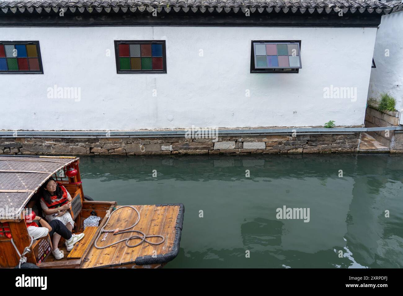 Suzhou, Cina - 11 giugno 2024: Una barca tradizionale cinese con due persone a bordo è ormeggiata accanto a una parete bianca con due finestre e una fontana di pietra Foto Stock