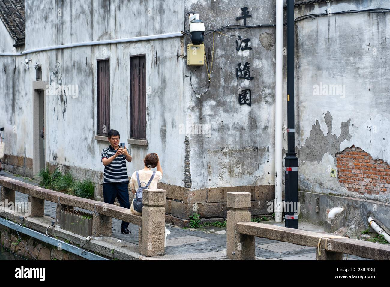 Suzhou, Cina - 11 giugno 2024: Un uomo scatta una foto di una donna seduta su una ringhiera in pietra accanto a un canale di fronte a un edificio bianco intempestivo con Chine Foto Stock