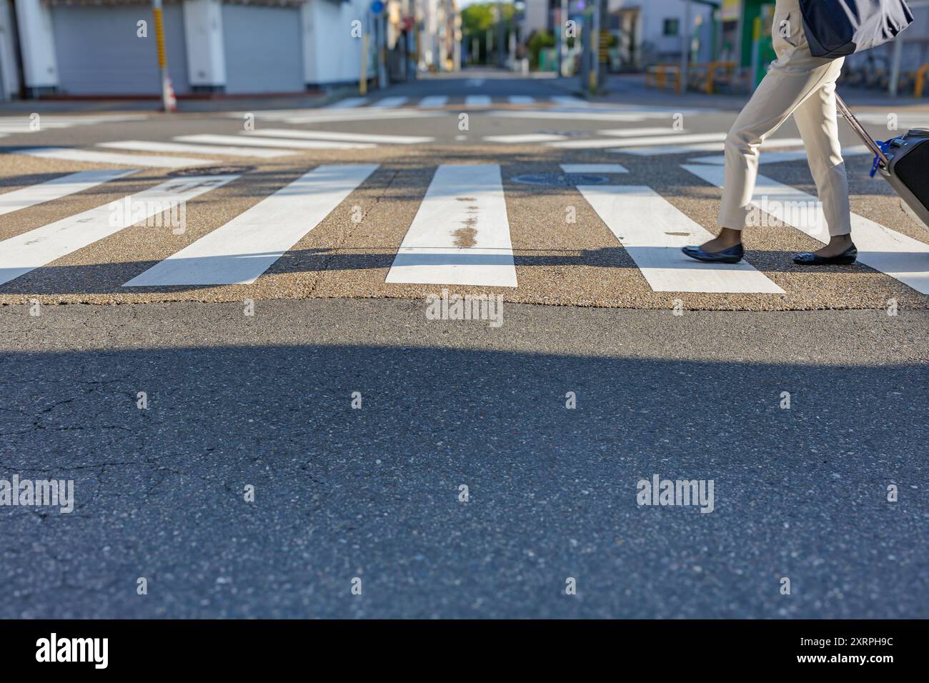 Una persona che tira una valigia con ruote attraversa un passaggio pedonale urbano vuoto. La luce del sole del mattino proietta lunghe ombre lungo la strada. Foto Stock