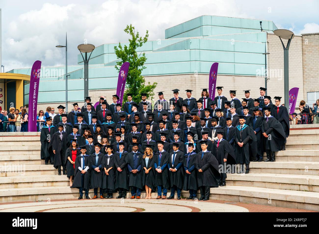 Gruppo di laureati fuori dalla Warwick University dopo la cerimonia di laurea, posando su alcuni passi preparandosi a fare il tradizionale lancio del berretto. Foto Stock