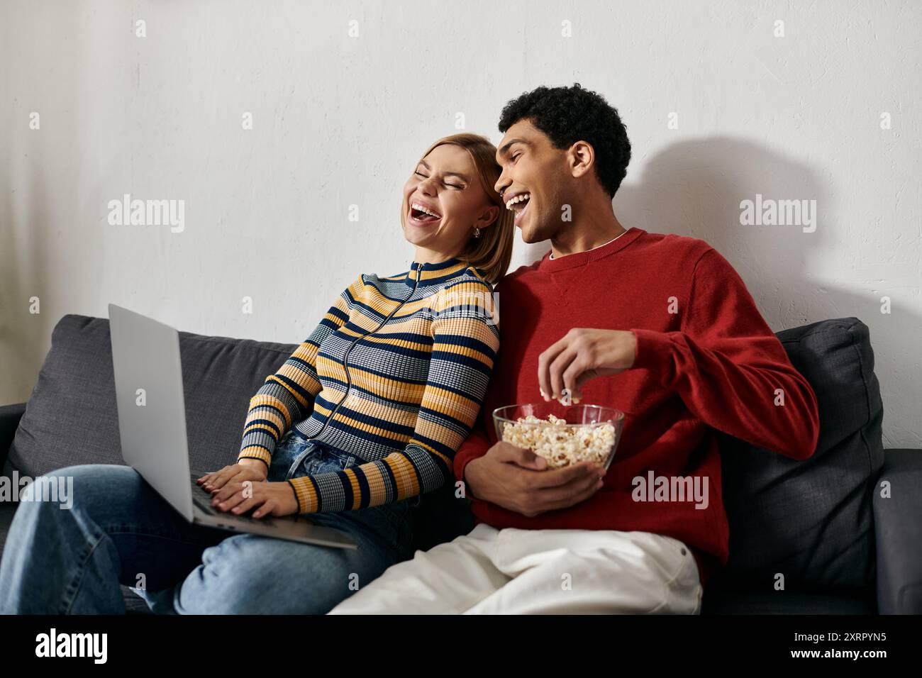 Una felice coppia interrazziale ride insieme mentre guardano un film su un computer portatile nel loro accogliente appartamento moderno. Foto Stock