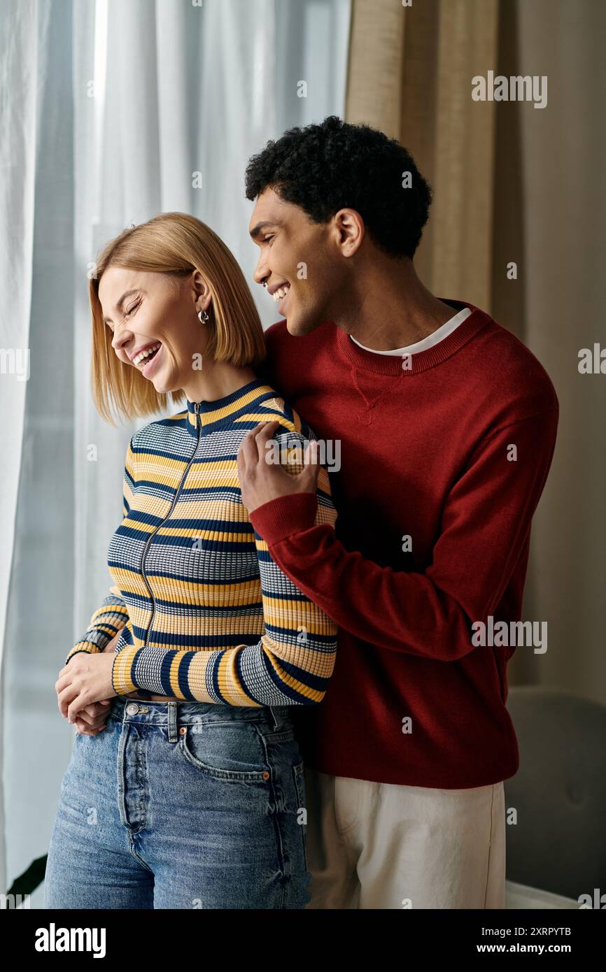Una coppia multiculturale, vestita con un abbigliamento casual, gode di un momento di risate insieme in un moderno ambiente di appartamenti. Foto Stock
