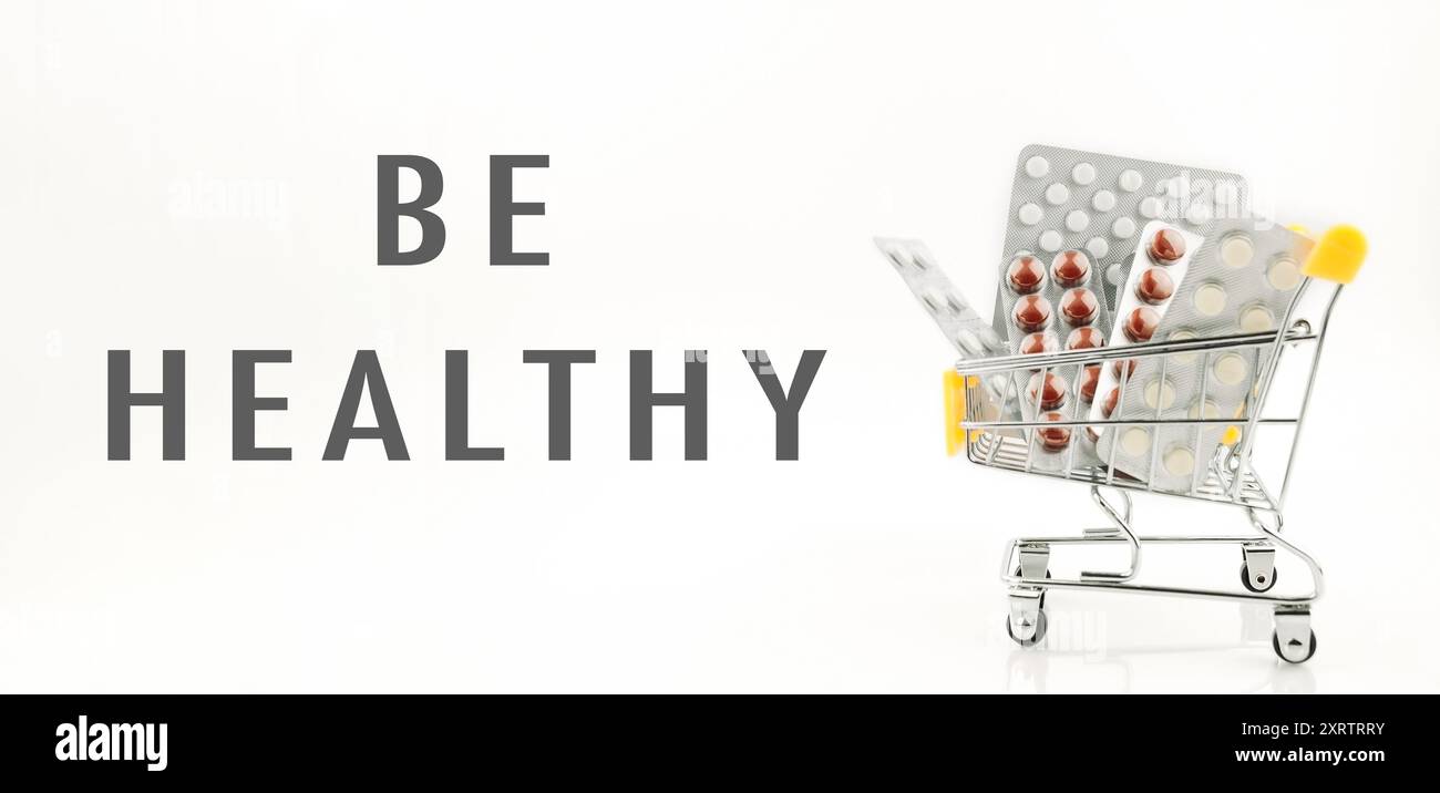 Un carrello pieno di pillole con le parole essere sano scritto sotto di esso. Concetto di prendersi cura della propria salute consumando vitamine e integratori Foto Stock