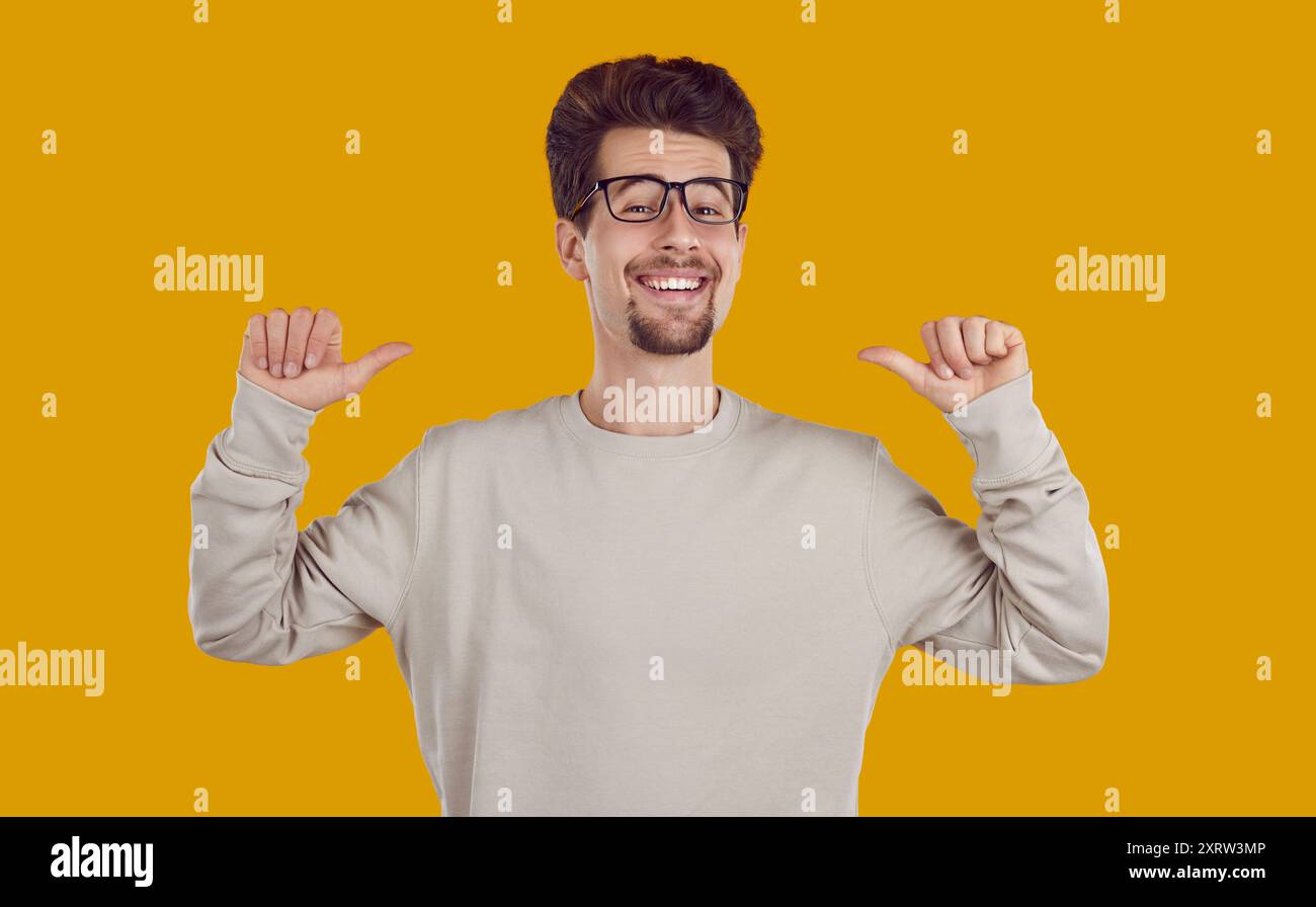 Sorridente sorridente felice uomo barbuto con gli occhiali che puntano i pollici a se stesso su sfondo giallo. Foto Stock
