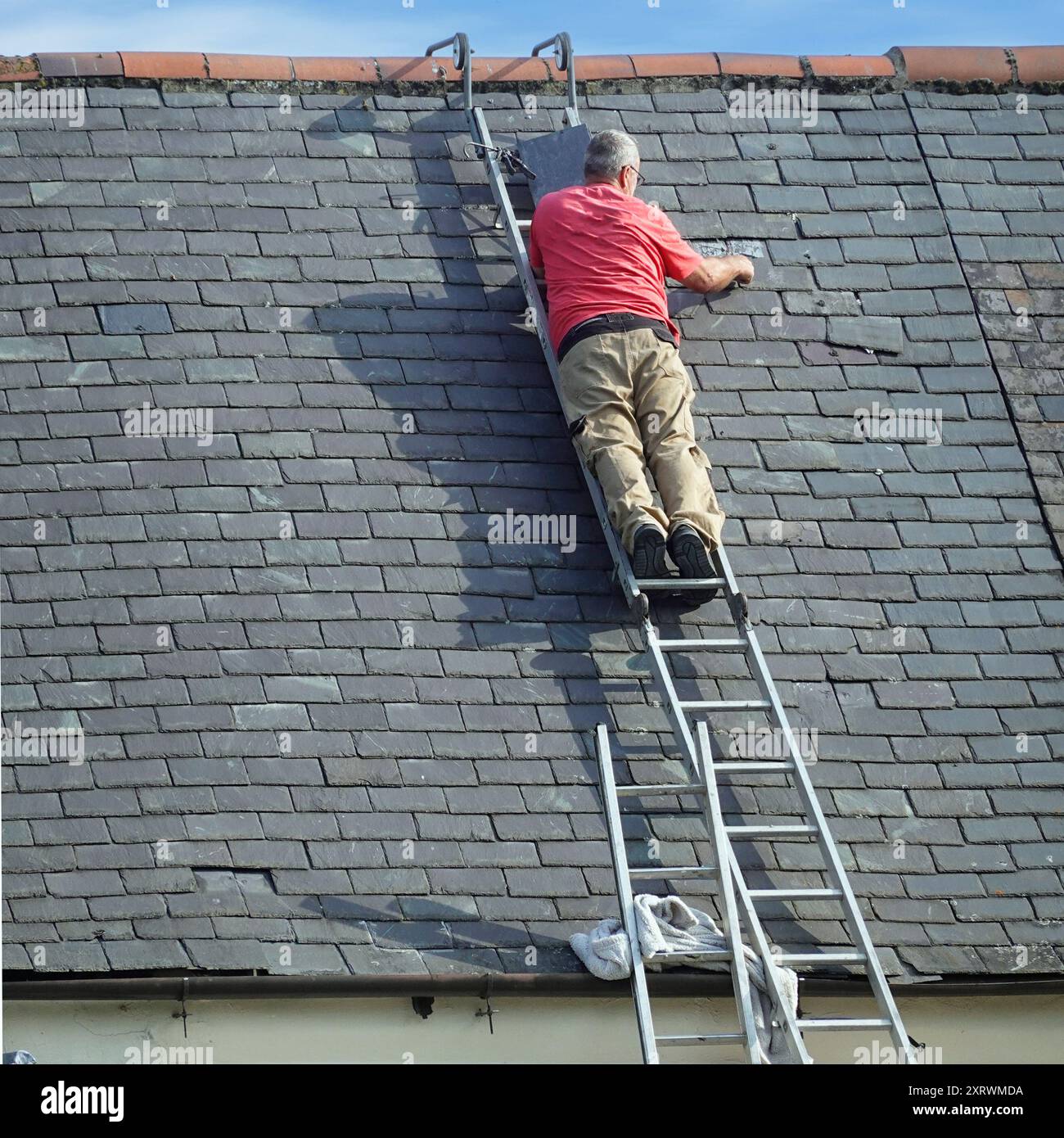 Commerciante con vista posteriore che lavora in posizione apparentemente precaria sostituendo ardelle danneggiate selezionate sul tetto inclinato della proprietà nella contea di Durham, Inghilterra, Regno Unito Foto Stock