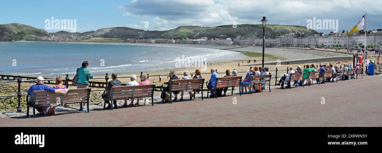 Vista sul retro, turisti adulti seduti su una lunga fila di panchine, ampia passeggiata panoramica verso Little orme Llandudno Conwy Clwyd, Galles del Nord, Regno Unito Foto Stock