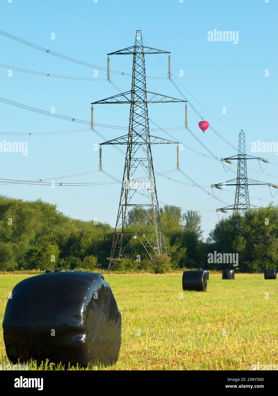 Piloni e pallone di passaggio a Kennington Meadows. Sono sempre stato affascinato dai nostri piloni britannici - quelle che in gran parte del mondo sarebbero chiamate torri di trasmissione. Marciando attraverso il paesaggio, hanno un impatto visivo così forte e sorprendente. Si trova a Kennington Meadows, tra Oxford e Abingdon, e una mongolfiera si aggira maestosamente nelle prime ore del mattino. Ma quali sono esattamente le misteriose luccicanti borse nere? Sfortunatamente, non sono escrementi alieni giganti. In realtà contengono insilato di balle per l'alimentazione del bestiame; per prima cosa, prendete il tradizionale rotolo di fieno e avvolgetelo in plastica A. Foto Stock