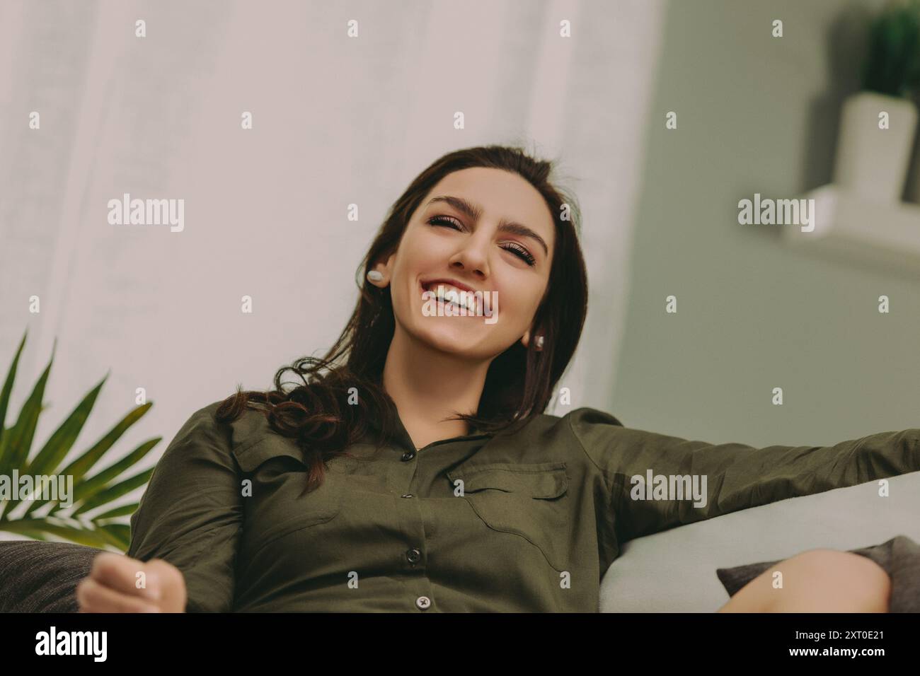 La donna con i capelli scuri e lisci sorride in grande mentre si rilassa su un divano moderno e chiaro. Vestita in modo informale con un top verde e pantaloncini bianchi, la sua espressione Foto Stock