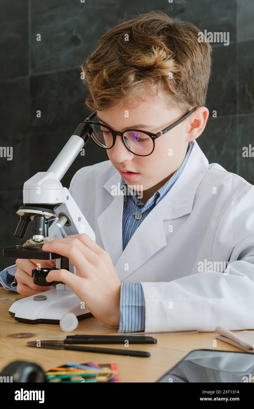 Preadolescente che indossa una provetta per l'esame con il microscopio mentre siede a tavola e si prepara alla ricerca in laboratorio a scuola in classe Foto Stock