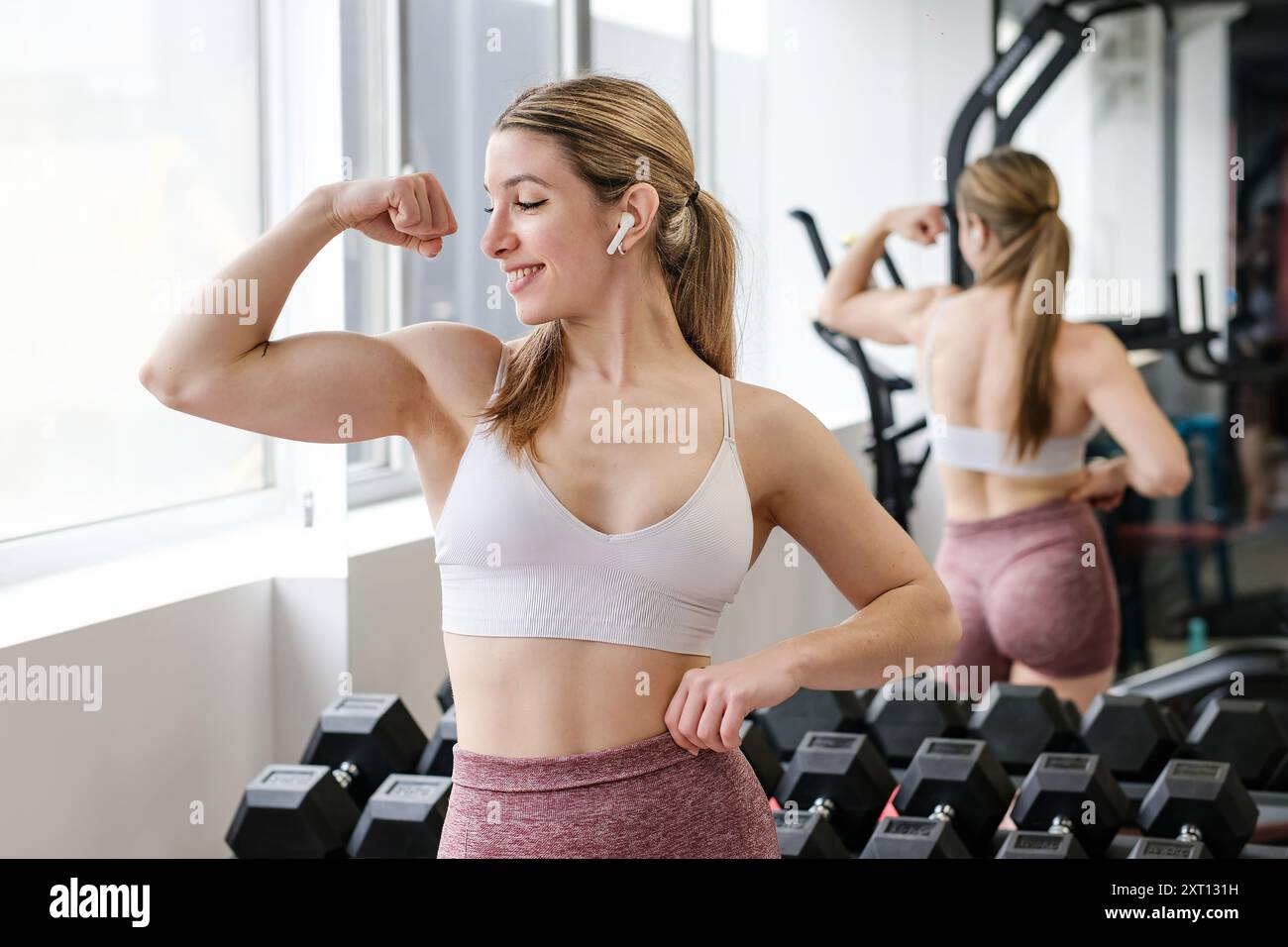 Una donna sorridente si flette il bicipite, ammirando il tono muscolare nello specchio della palestra, irradiando salute e sicurezza Foto Stock