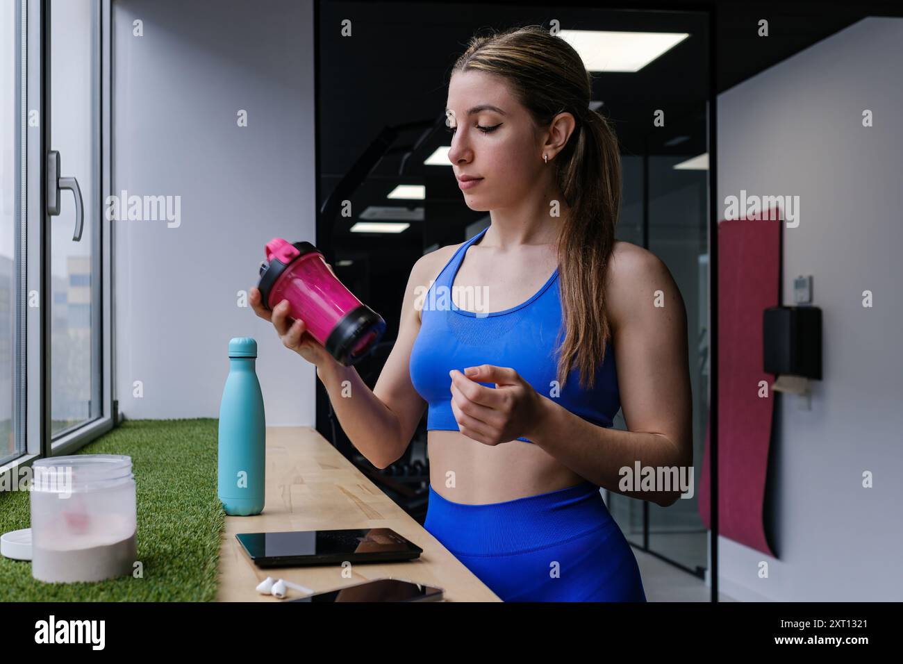 Un'atleta concentrata in un outfit sportivo blu coordinato mescola il suo drink post-allenamento con uno shaker portatile Foto Stock