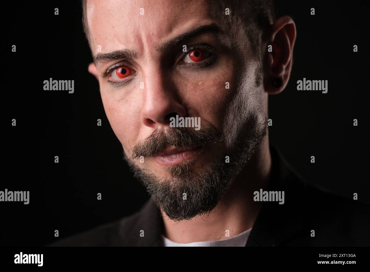 Ritratto ravvicinato di un uomo barbuto con un'espressione poderosa che indossa lenti a contatto rosse, illuminato da una luce laterale spettacolare che contrasta nettamente Foto Stock