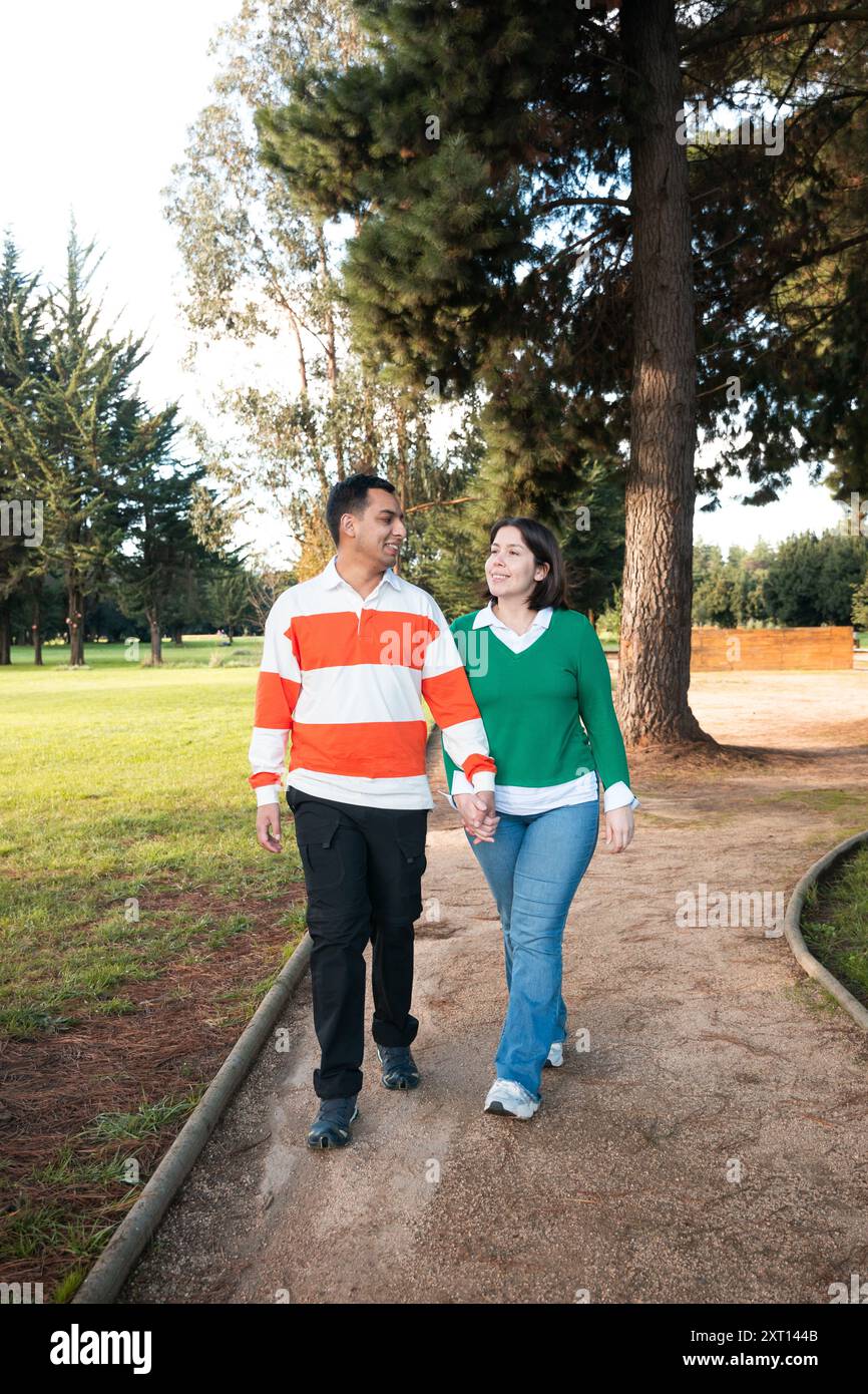 Una giovane coppia latina si gode una piacevole passeggiata attraverso un parco verdeggiante, condividendo un momento di connessione mentre camminano mano nella mano, con alberi alti nel Foto Stock