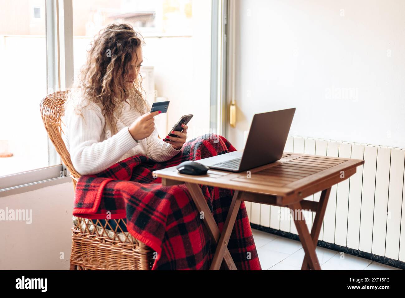 Giovane donna dai capelli ricci in maglione bianco seduto a tavola con il laptop mentre utilizza lo smartphone e la carta di credito per l'acquisto online via Internet Foto Stock