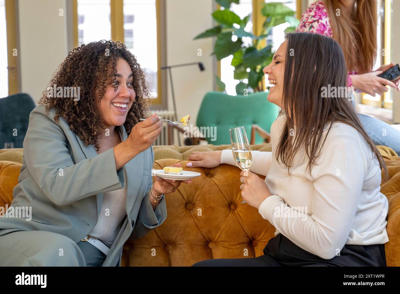 Due donne ridono e si godono cibo e bevande insieme su un comodo divano in una camera ben illuminata. Panc05700 Copyright: XConnectxImagesx Foto Stock