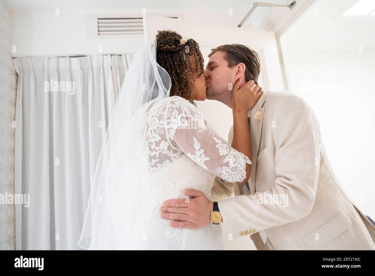 La sposa e lo sposo condividono un bacio appassionato il giorno del loro matrimonio, con lo sposo in abito beige e la sposa con un abito e un velo in pizzo bianco. panc059 Foto Stock