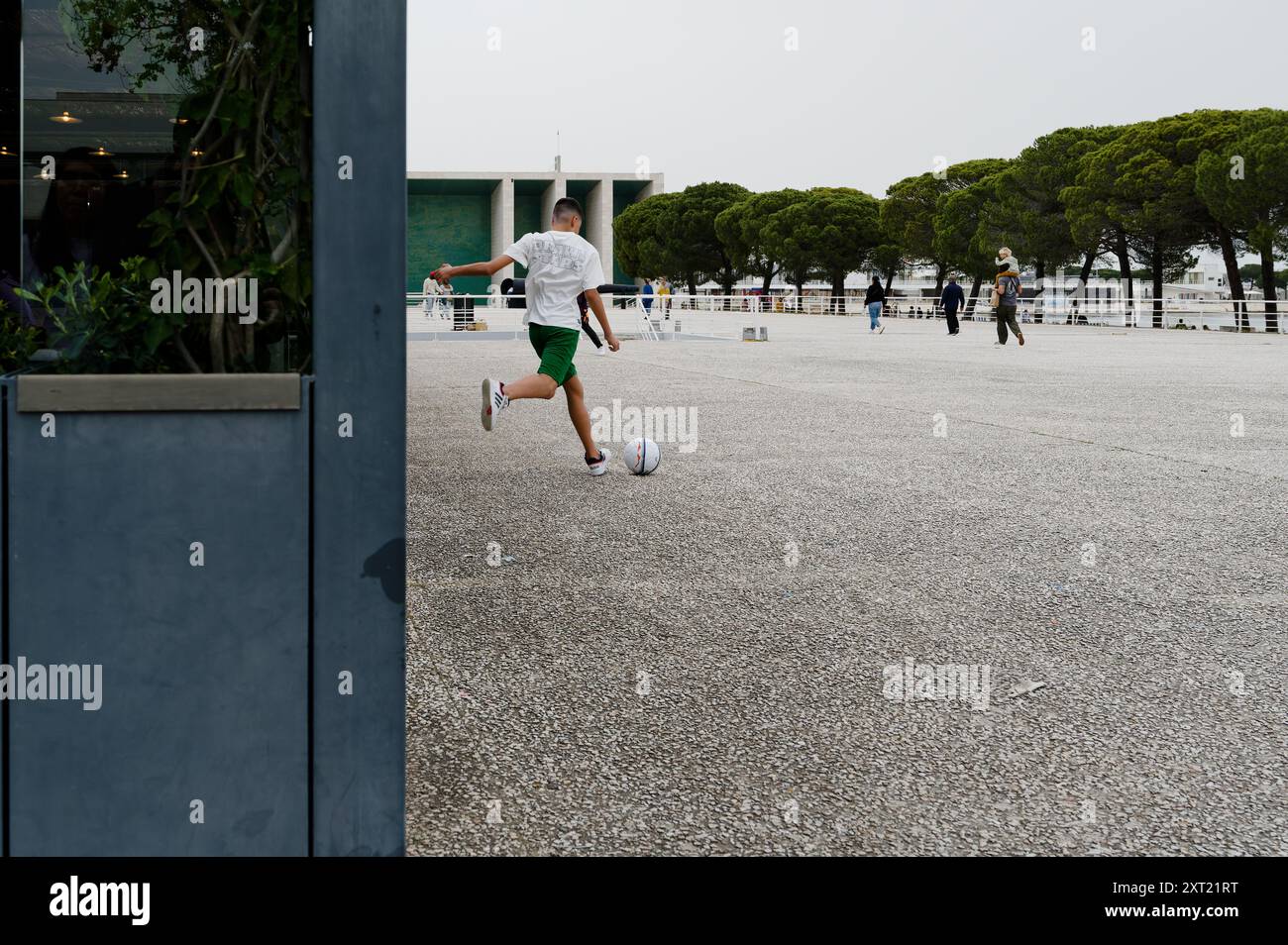 Un adolescente calci un pallone da calcio in una spaziosa piazza di Lisbona, circondata da architettura moderna e vegetazione Foto Stock