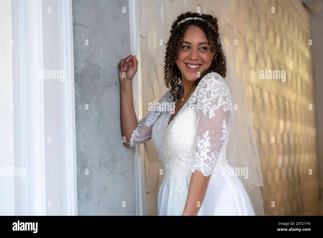 Sposa radiosa in un abito da sposa in pizzo con un sorriso travolgente che si erge su una parete testurizzata bianca. Panc05962 Copyright: XConnectxImagesx Foto Stock