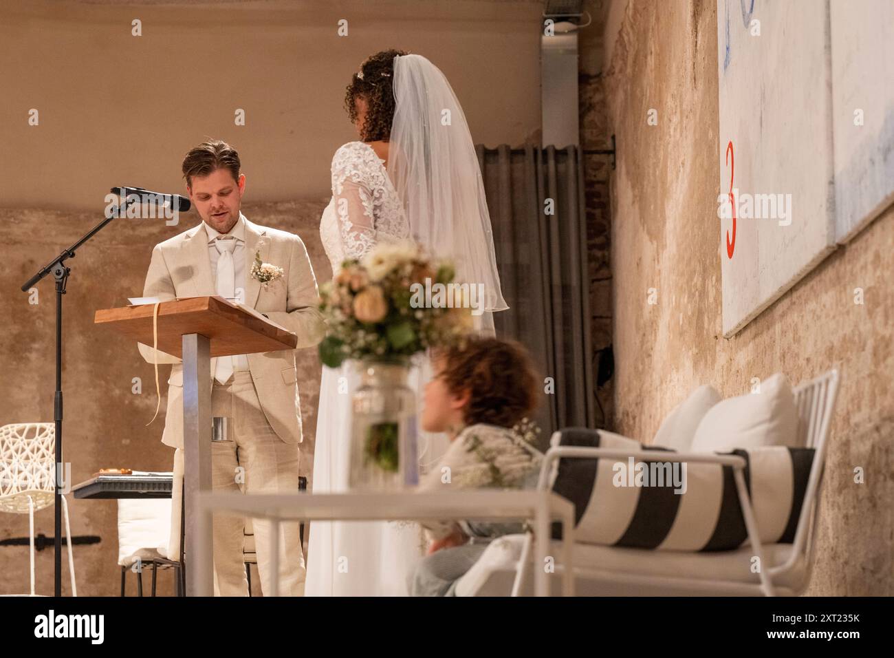 Uno sposo con un vestito di colore chiaro si erge su un podio con un microfono, guardando una sposa con un vestito bianco e un velo durante una cerimonia nuziale in ruggine Foto Stock