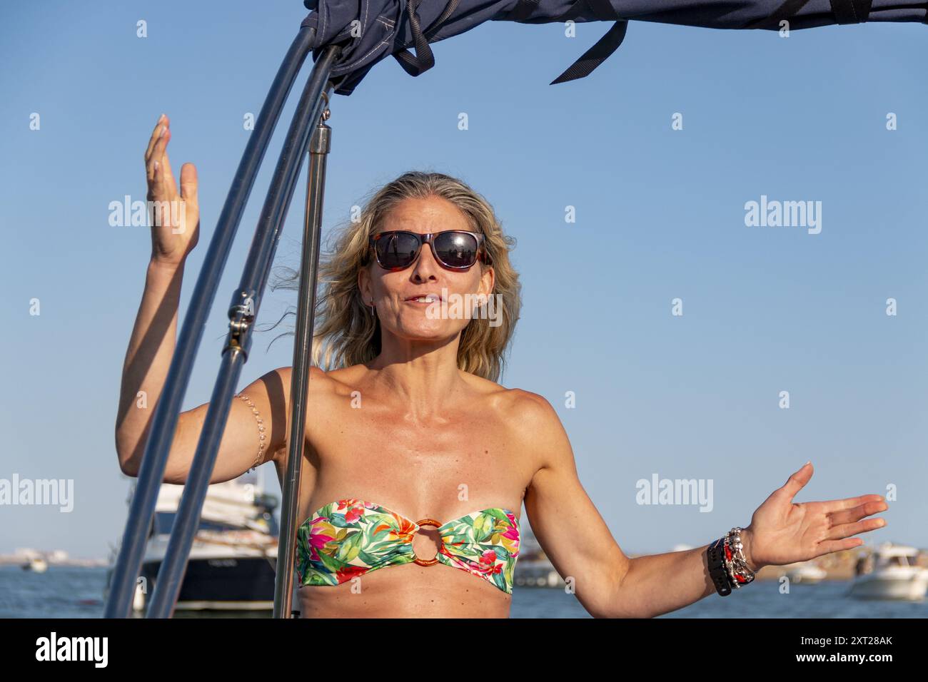 Donna bionda con capelli sciolti e occhiali da sole si appoggia sul lato di una barca sopra le onde e il mare blu Foto Stock