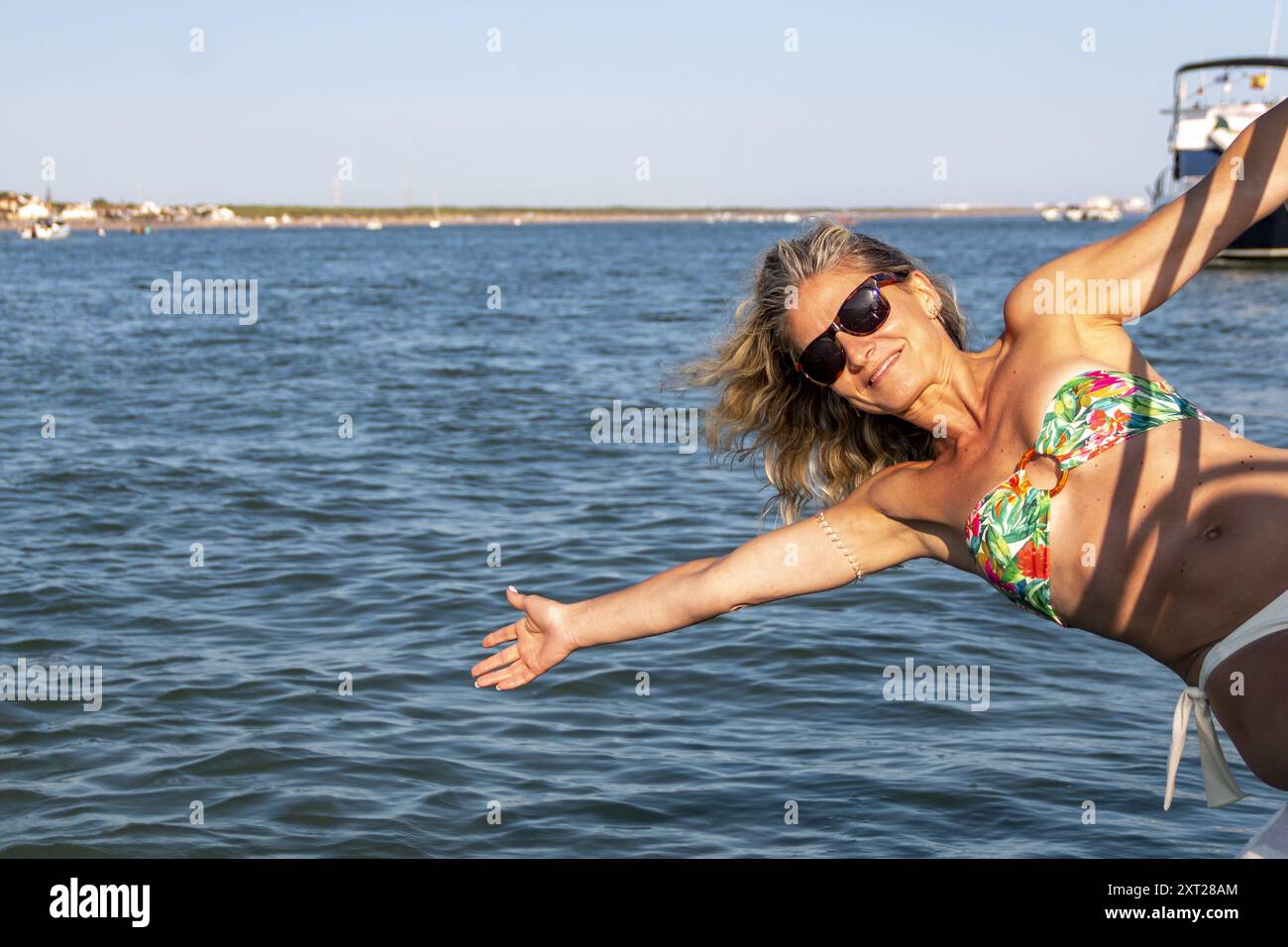 Donna bionda con capelli sciolti e occhiali da sole si appoggia sul lato di una barca sopra le onde e il mare blu Foto Stock