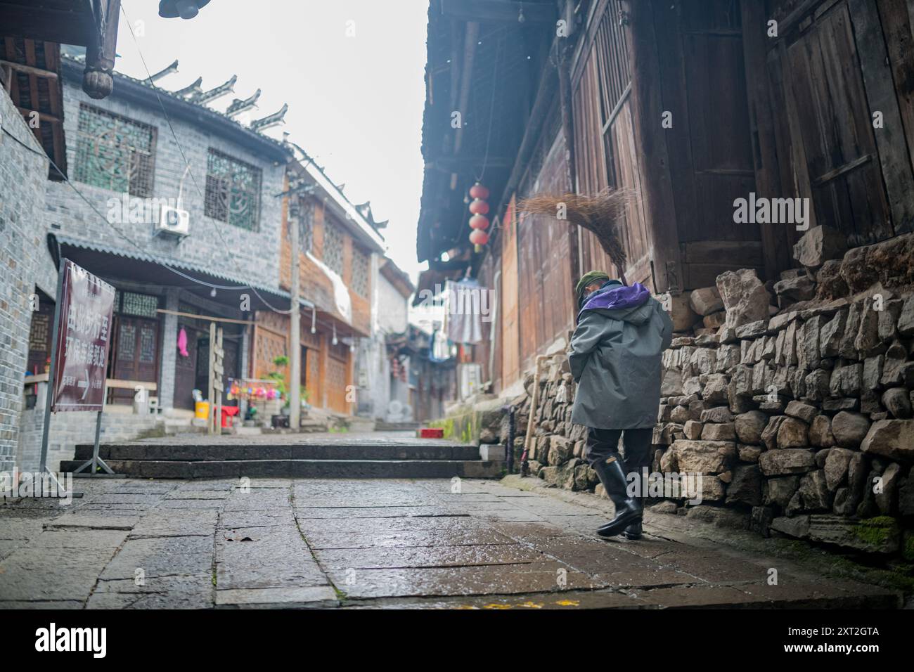 Un uomo anziano che spazia su strade di ciottoli bagnate in un tradizionale villaggio cinese, circondato da un'architettura in legno e pietra. Foto Stock
