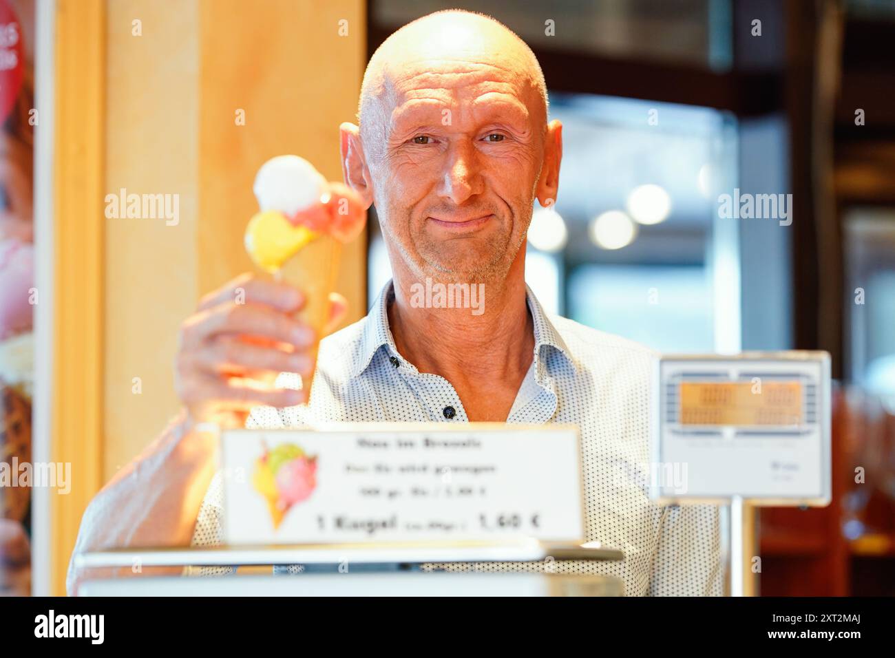 PRODUZIONE - 13 agosto 2024, Baden-Württemberg, Gaggenau: L'operatore Michael Böhmer mette un gelato in un cono sulla bilancia al Café Brezels. Al Café Brezels, nel centro di Gaggenau, il gelato non viene più caricato dalla paletta ma dal grammo. I clienti pagano due euro per 100 grammi di gelato e il prezzo viene adeguato di conseguenza per quantità minori. Foto: Uwe Anspach/dpa Foto Stock